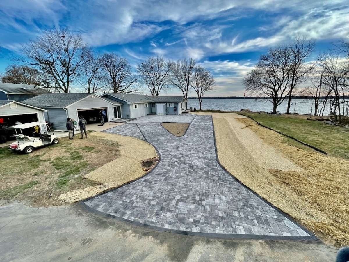 Driveway with gray pavers, tan gravel, and a gray garage next to a golf cart, trees, and water under a blue sky.