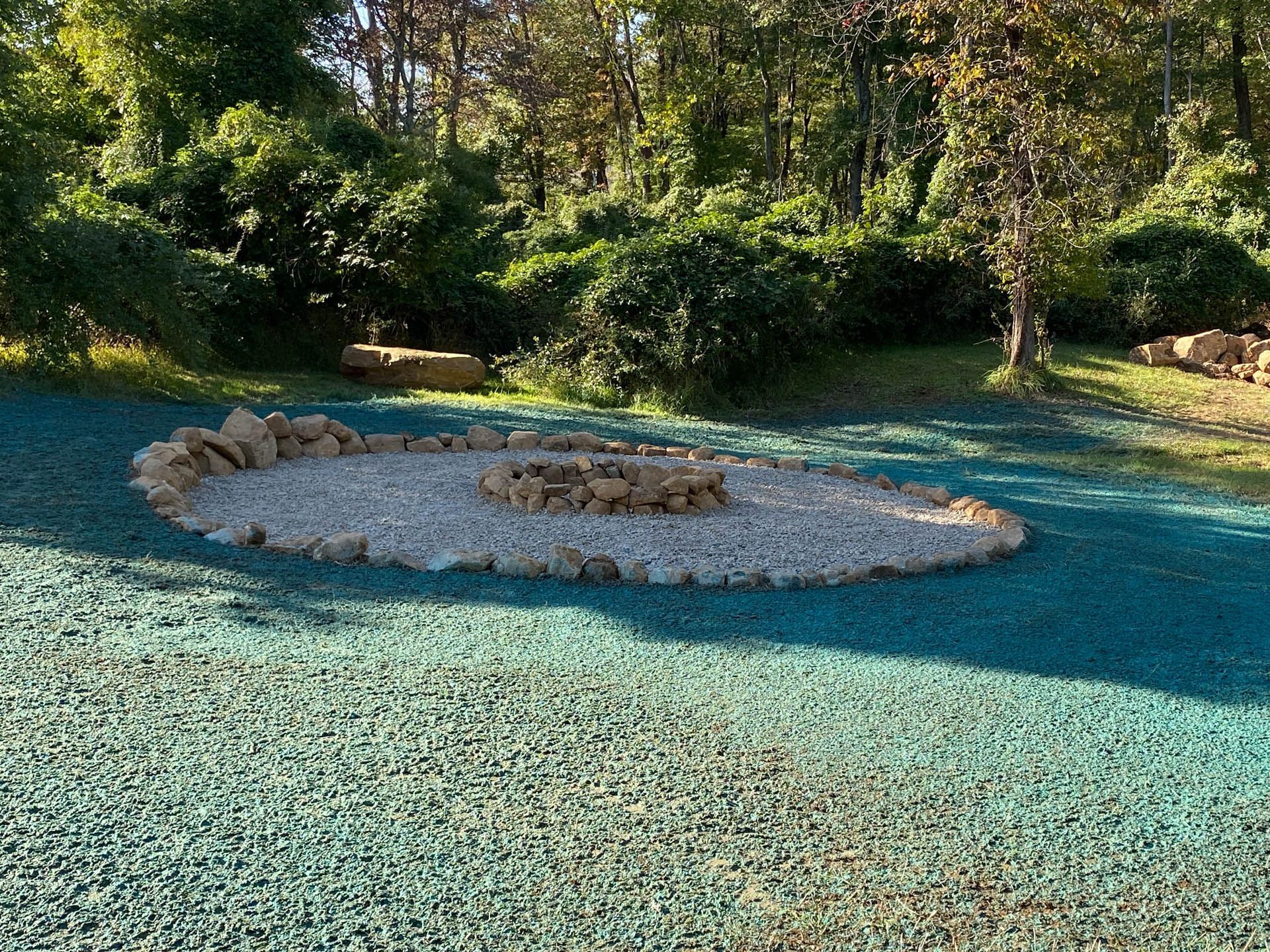 Oval fire pit with a stone border on a teal-colored rubber surface in a wooded area.