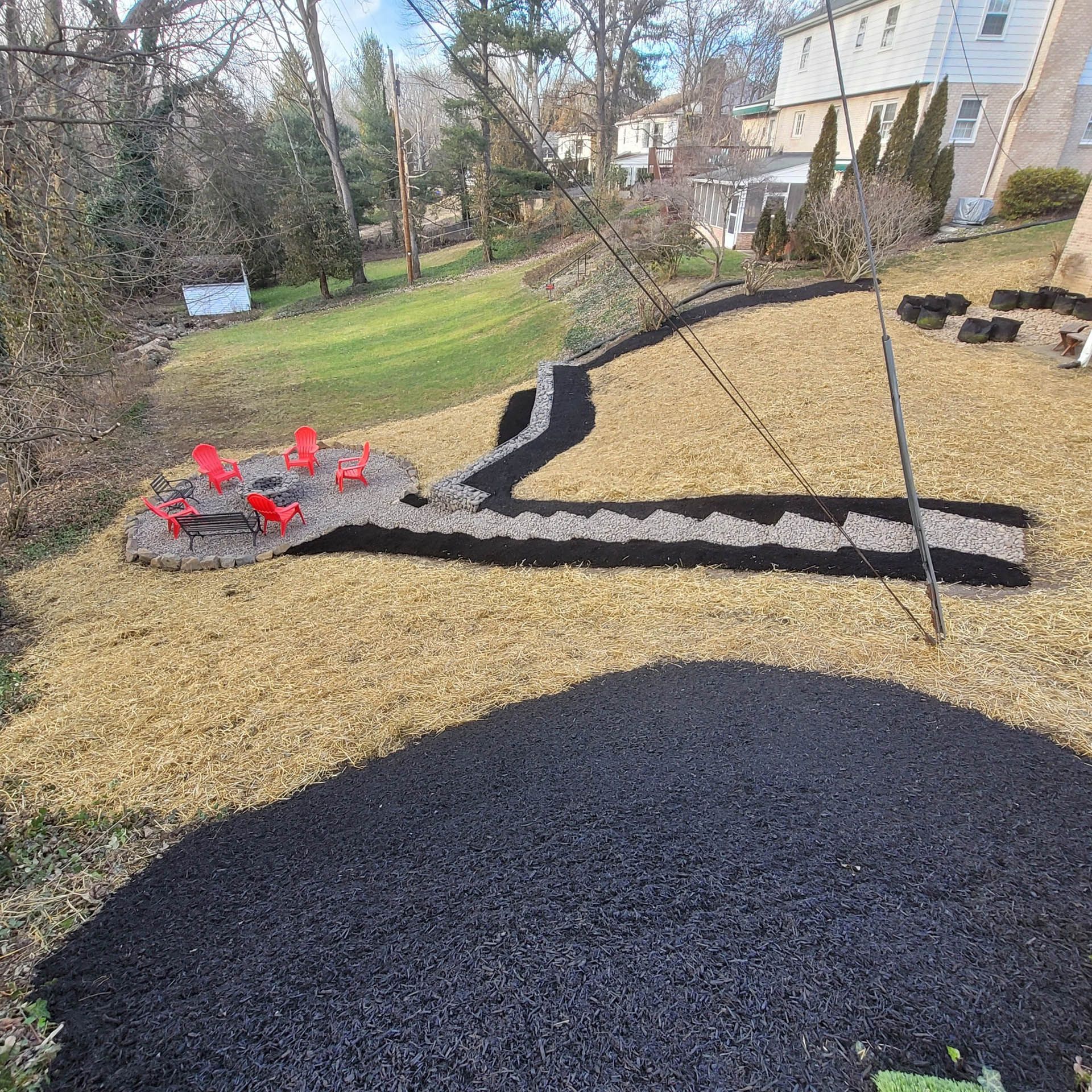 Backyard landscape with fire pit and dark mulch borders. Brown grass, red chairs, and a white house.