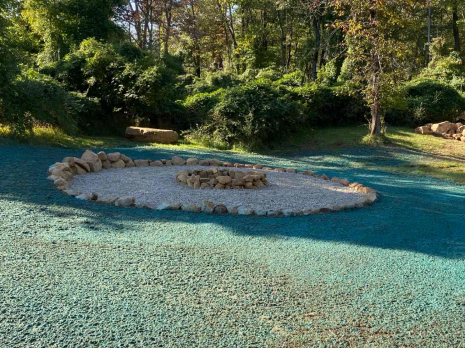 Fire pit ringed by rocks, on a gravel surface with artificial turf in a yard, trees in the background.