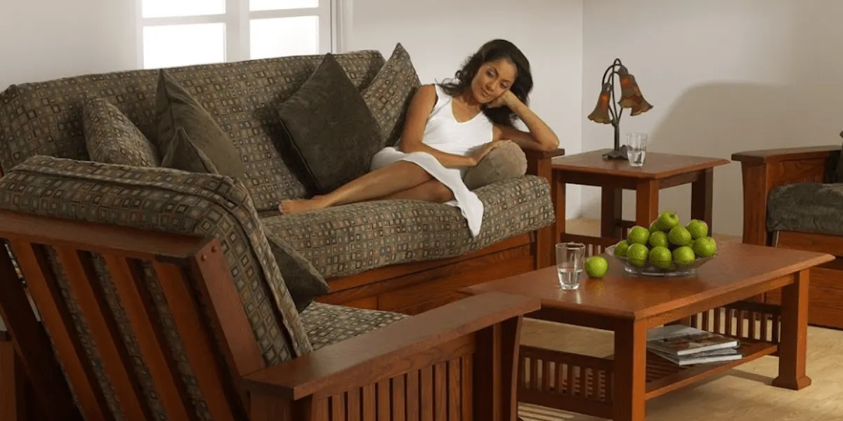 Woman relaxing on a brown futon in a living room, with a table, lamp, and fruit bowl.
