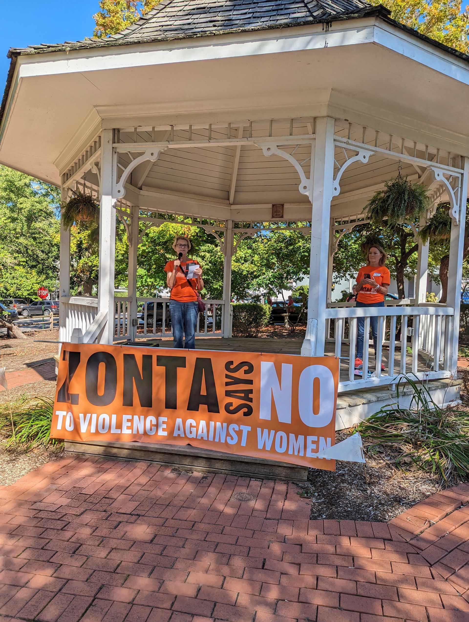 Two people in orange shirts stand in a gazebo holding a banner that reads