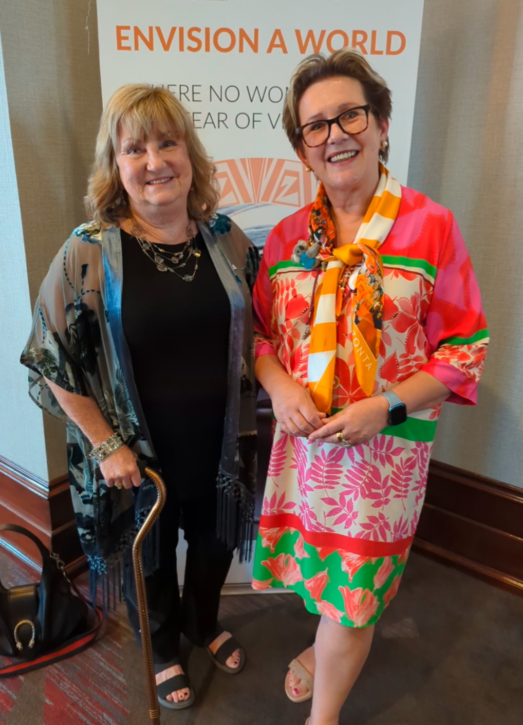 Two women smiling, posing beside a banner. One holds a cane, wearing black; the other wears a colorful dress.
