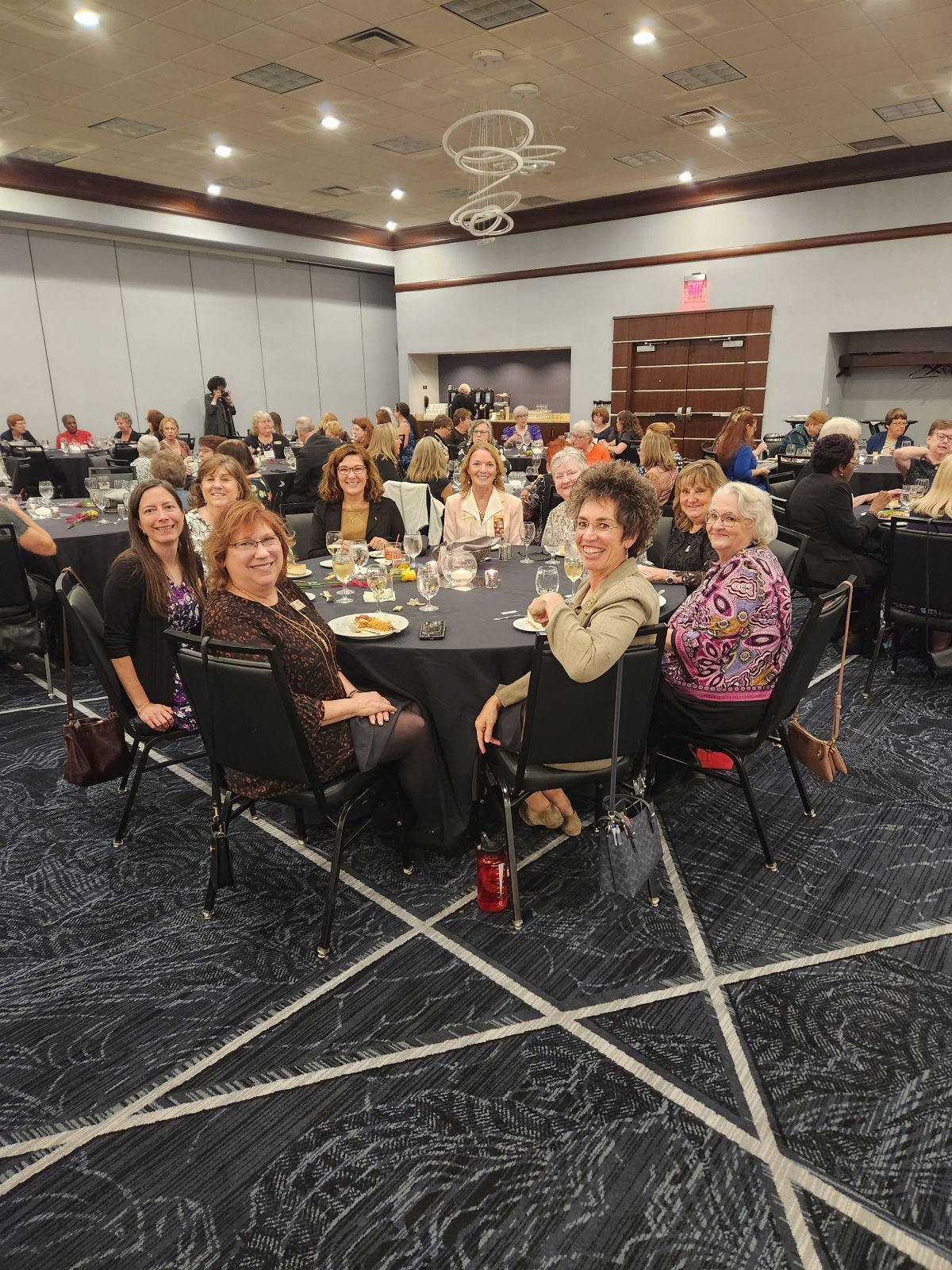 Attendees seated at round tables in a large event room. Several women smile, some hold drinks.