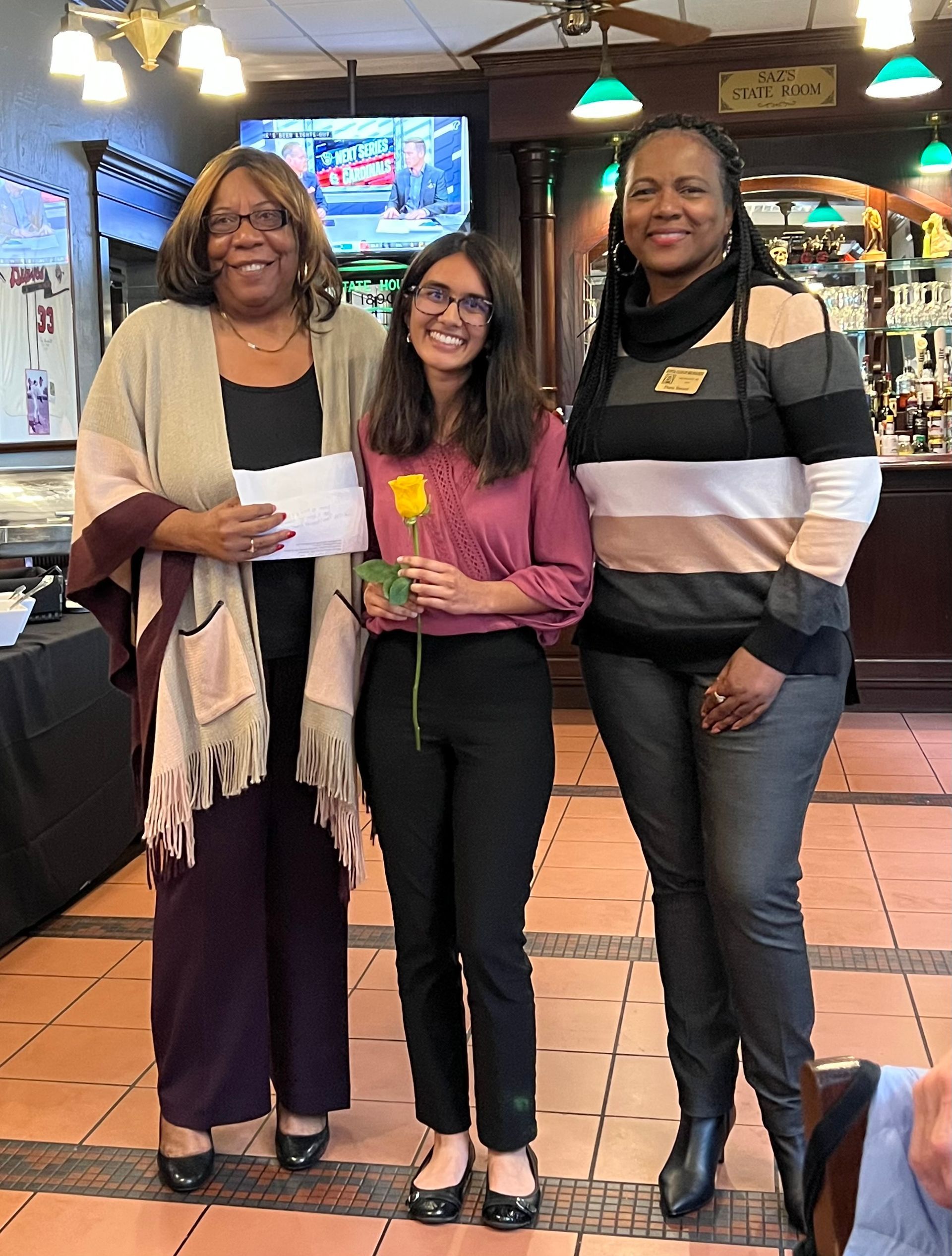 Three women smiling, two holding a check and flower, standing indoors in front of a bar.