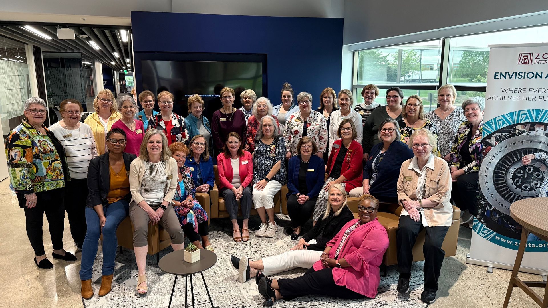 Group of women in a modern office setting, smiling for a photo. Some seated, others standing.