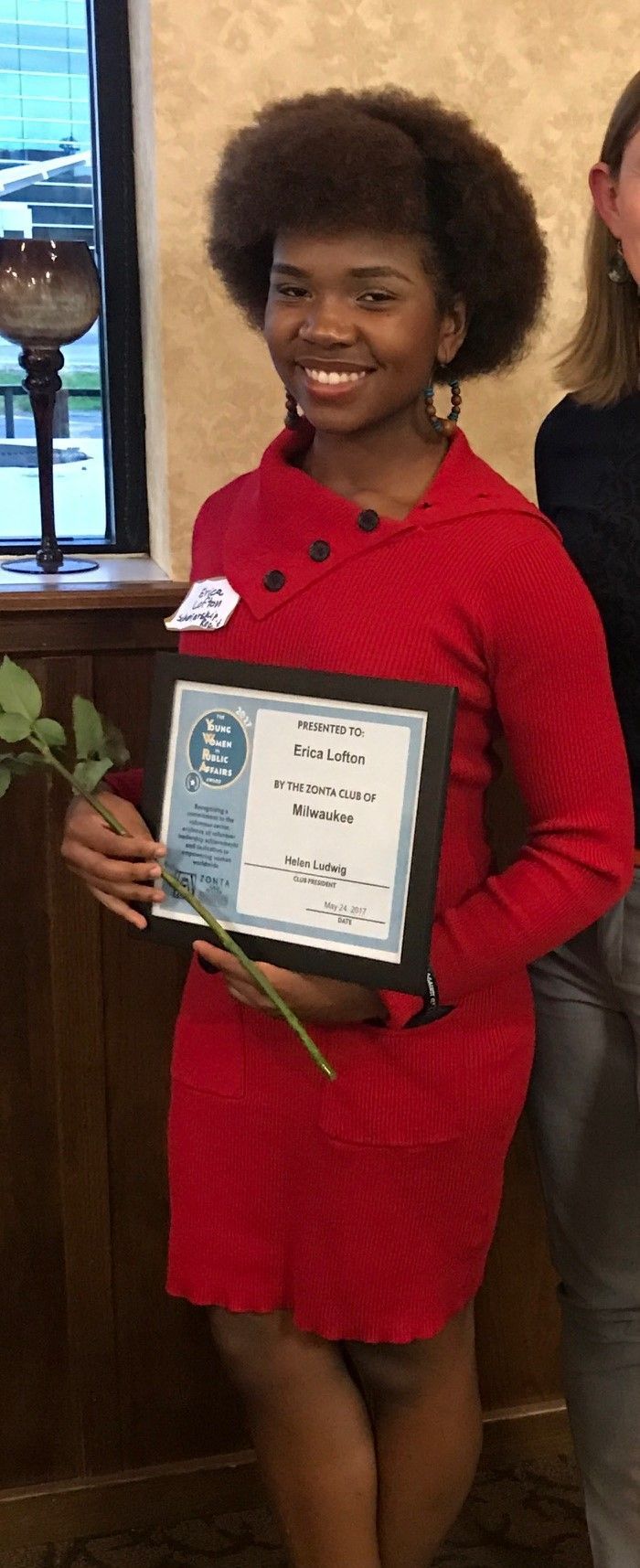 Young Black woman smiles, holding an award. She wears a red dress and has large, natural hair.