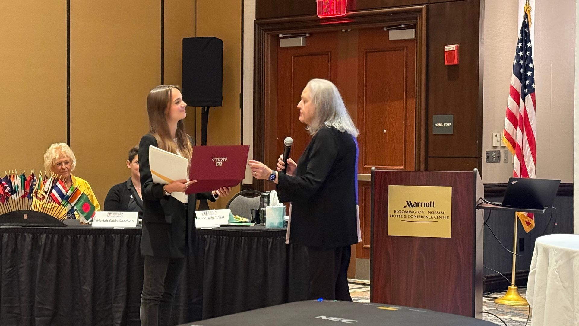 Woman presenting award to another woman at a podium. American flag in background.