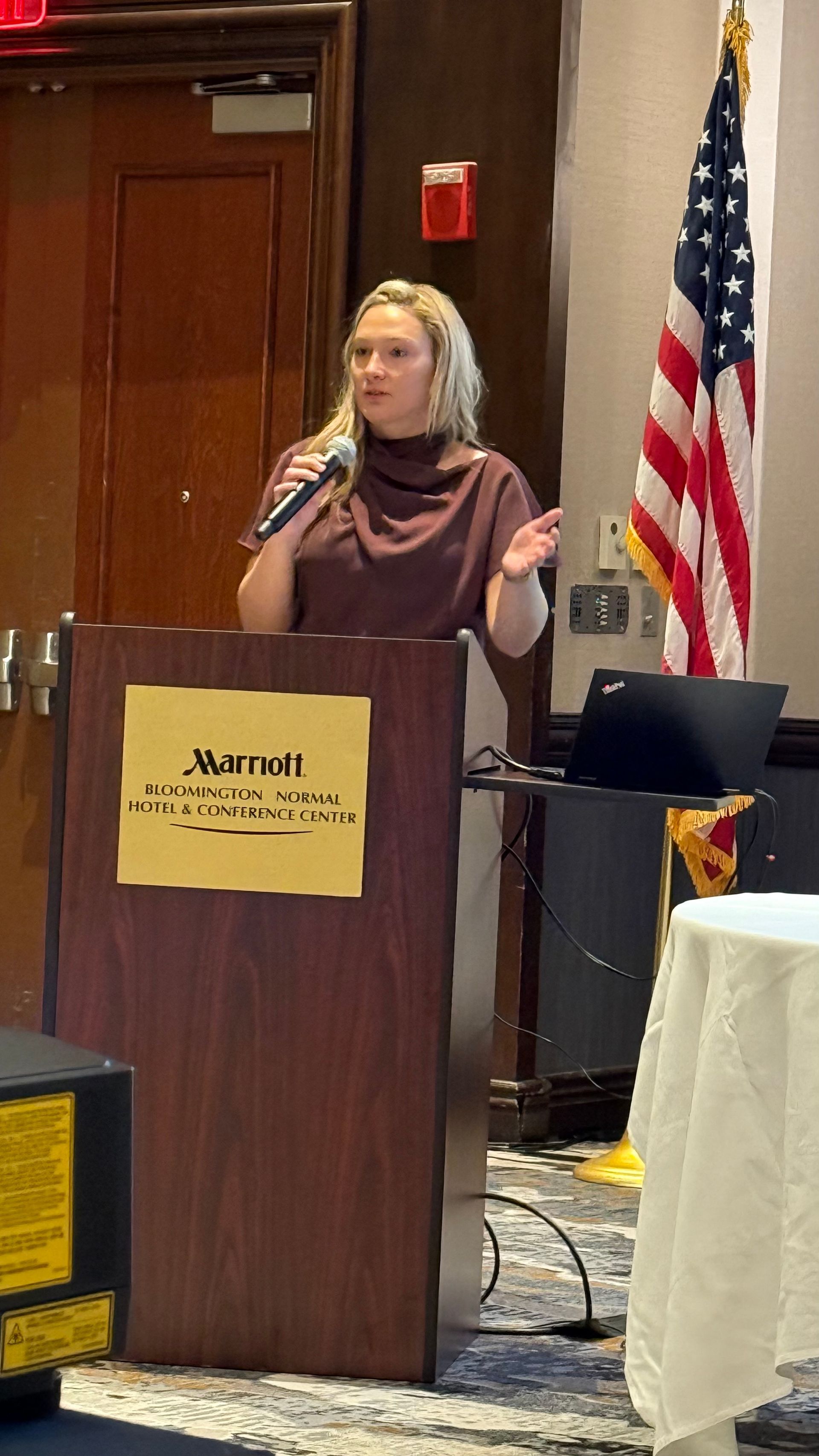 Woman speaking at a Marriott podium with US flag in the background. She wears a brown top and raises her arms.