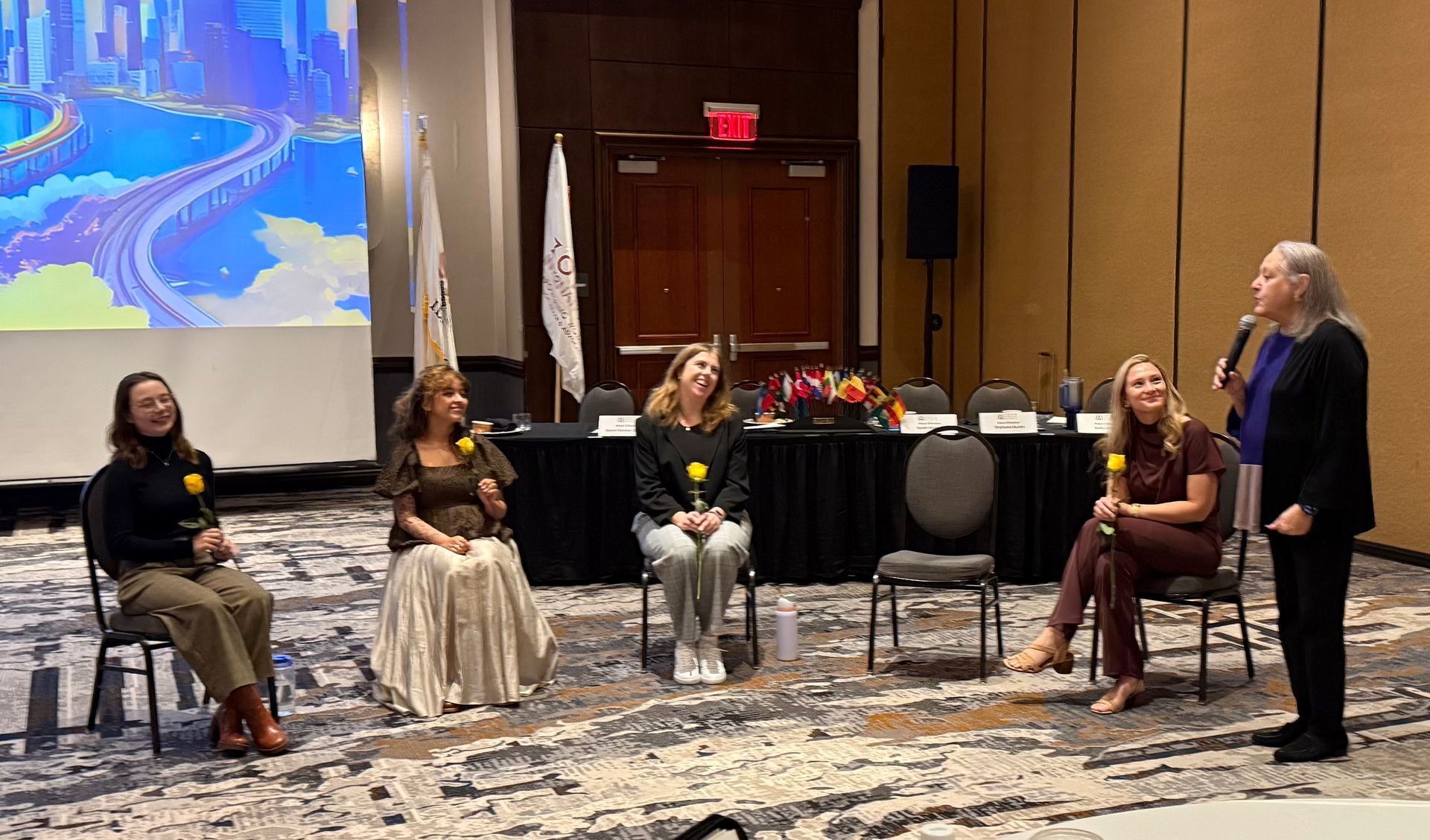 Five people, including one with a microphone, seated on chairs at a conference. They hold flowers.