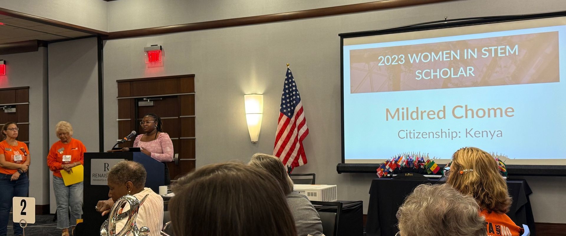 Woman speaking at podium at a conference with a projector screen. The screen says