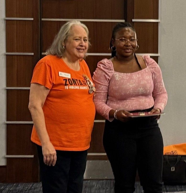 Two women smiling, one in orange shirt, other in pink, holding certificate.
