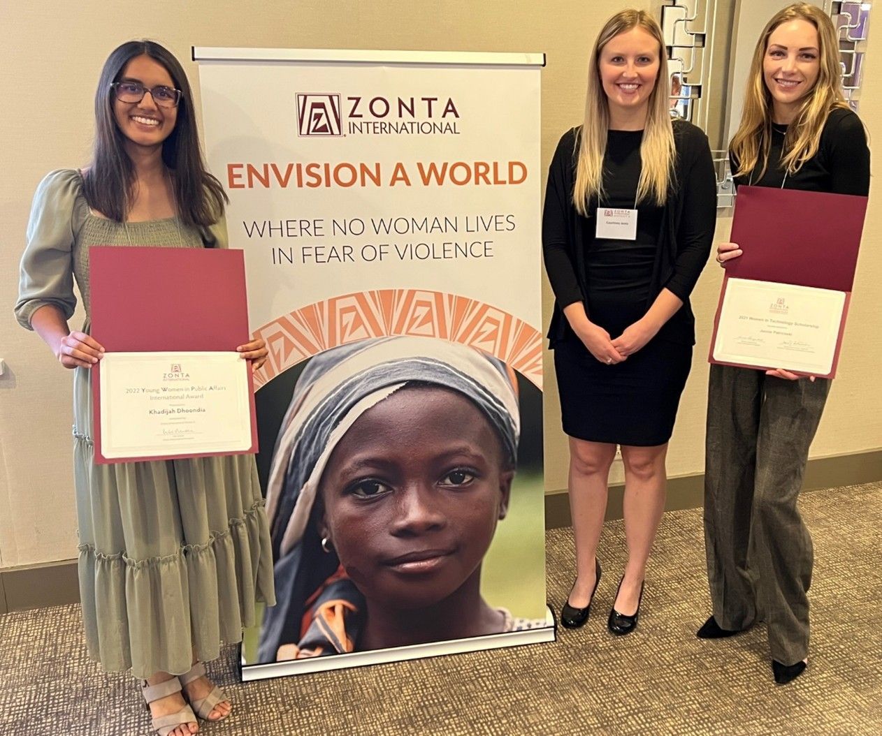 Three women stand with awards beside a Zonta International banner. The banner shows a child's face and text.
