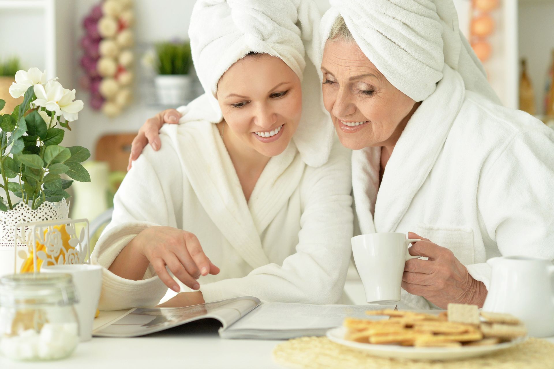 Two people in white robes and towels read a cookbook at a table with tea and cookies