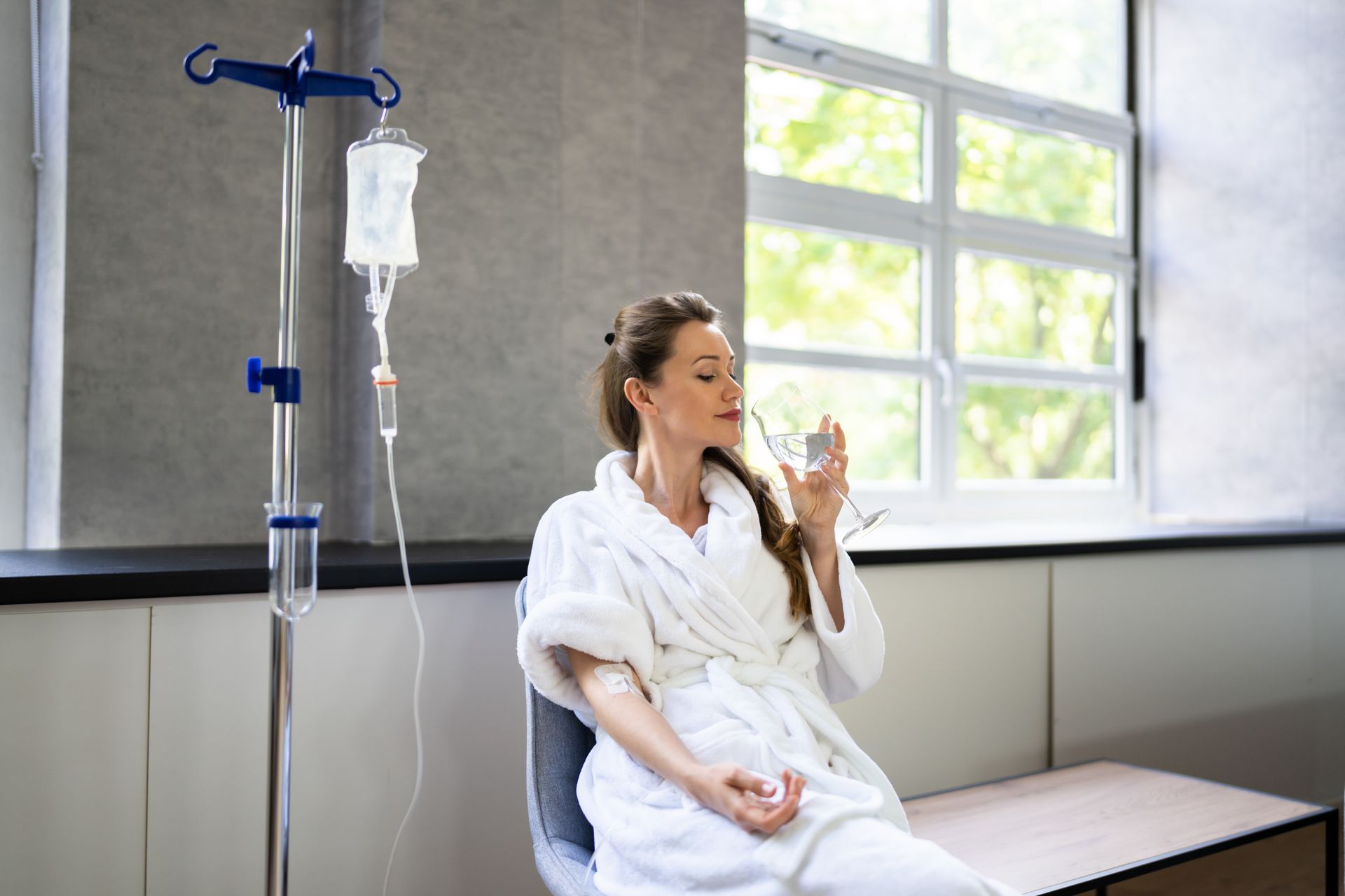 Woman in a white robe drinking water beside an IV stand in a bright hospital room