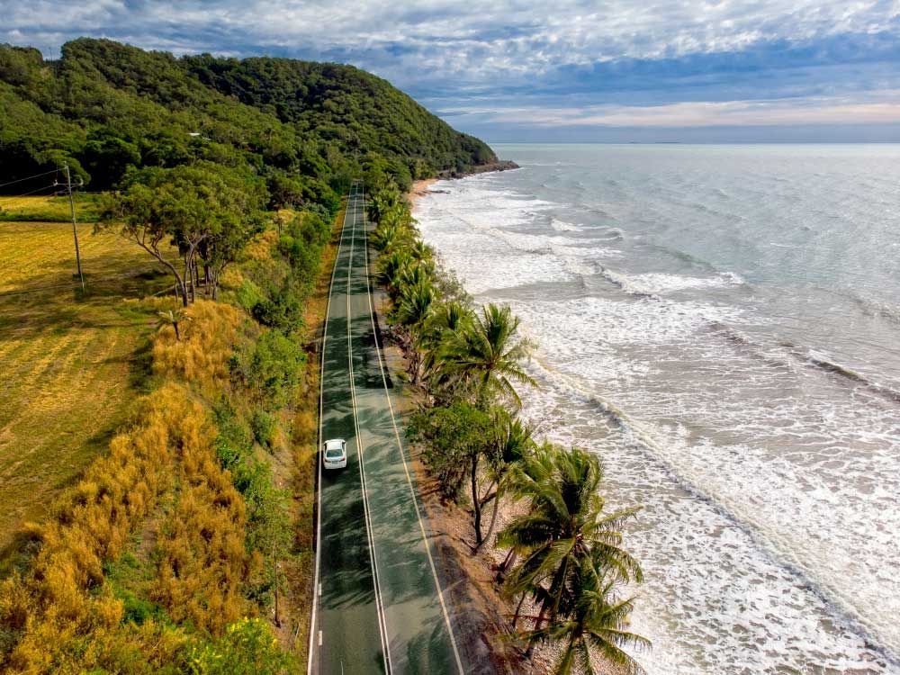An Aerial View Of A Car Driving Down A Road Next To The Ocean — Barron River Lawns In Mossman, QLD

