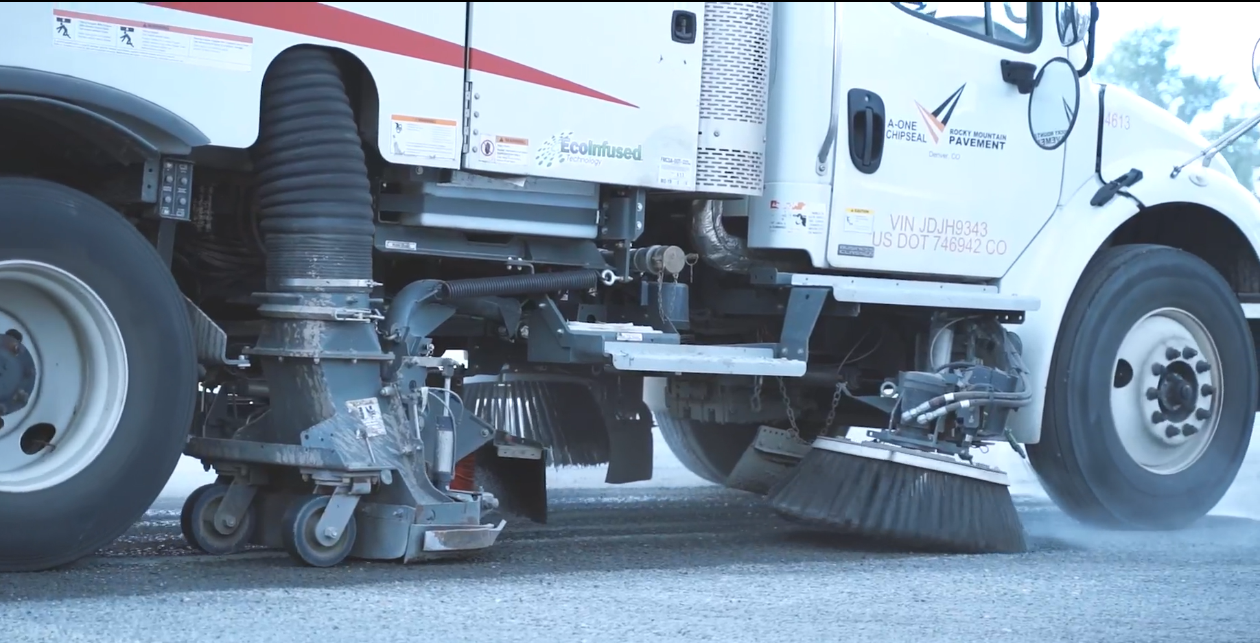Street sweeper cleaning a road with rotating brushes, wheels, and a white truck.