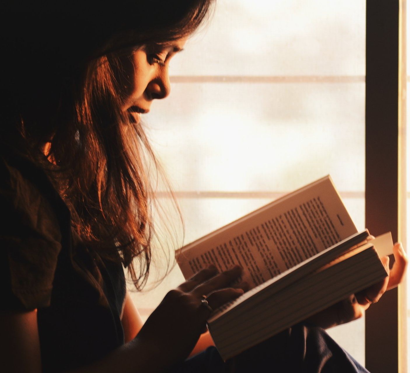 A woman is reading a book in front of a window, studying.