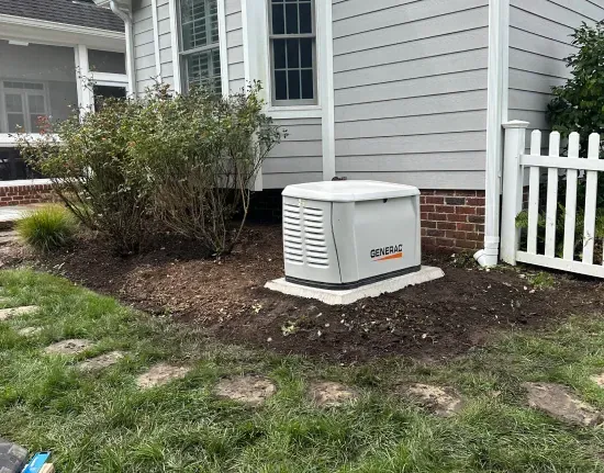 White home generator on a concrete pad beside a gray house and white fence, with mulch and shrubs nearby