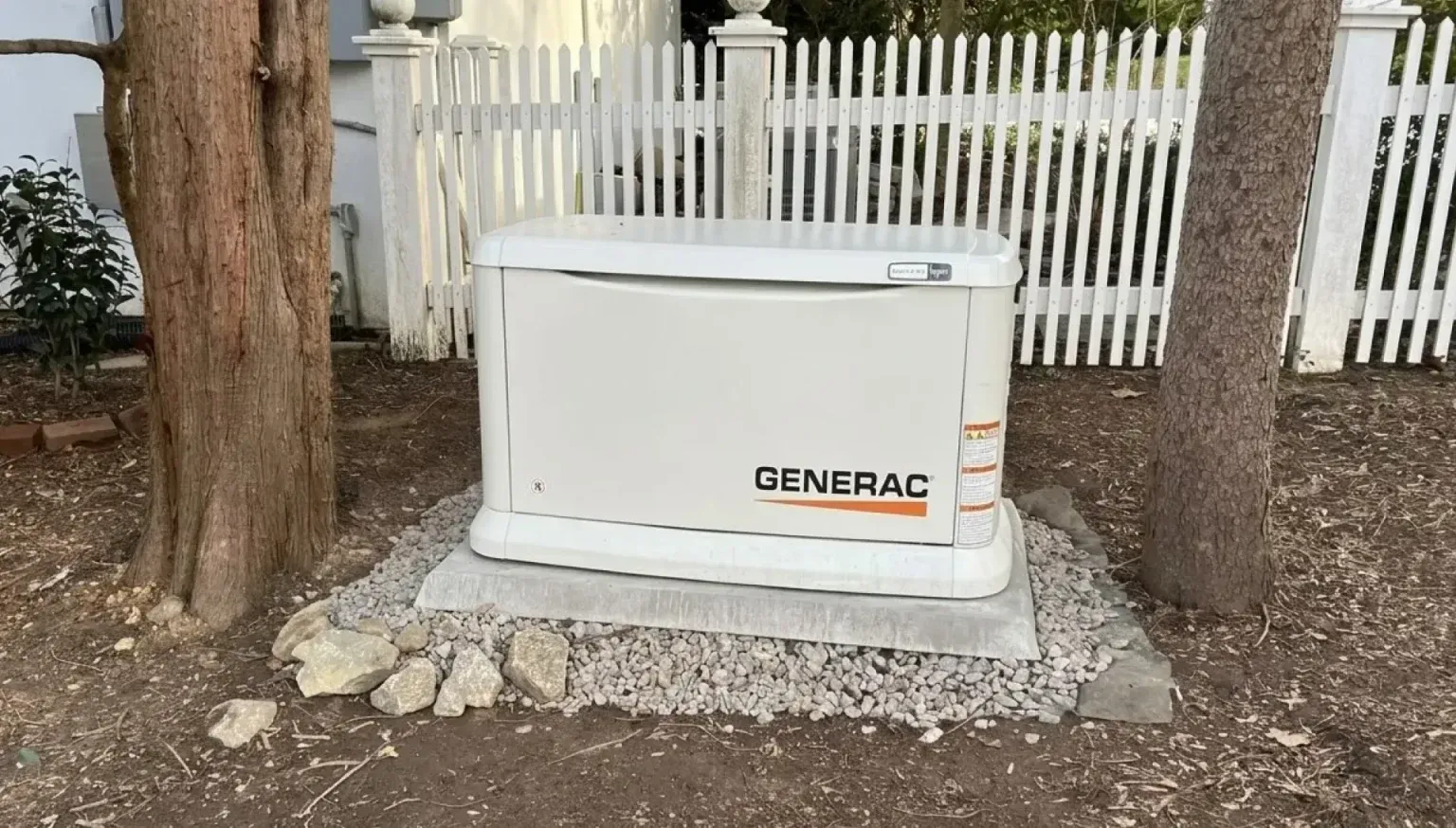 White GENERAC generator beside a tree and white picket fence, set on a gravel base outdoors
