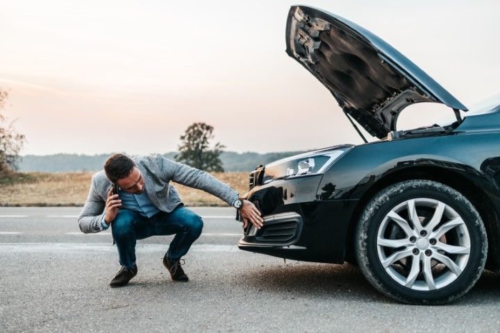 Man crouching beside a black car with the hood open, inspecting the front bumper on a roadside.