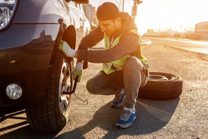 Person crouches by a car’s front wheel, using a wrench beside a spare tire on a sunlit roadside.