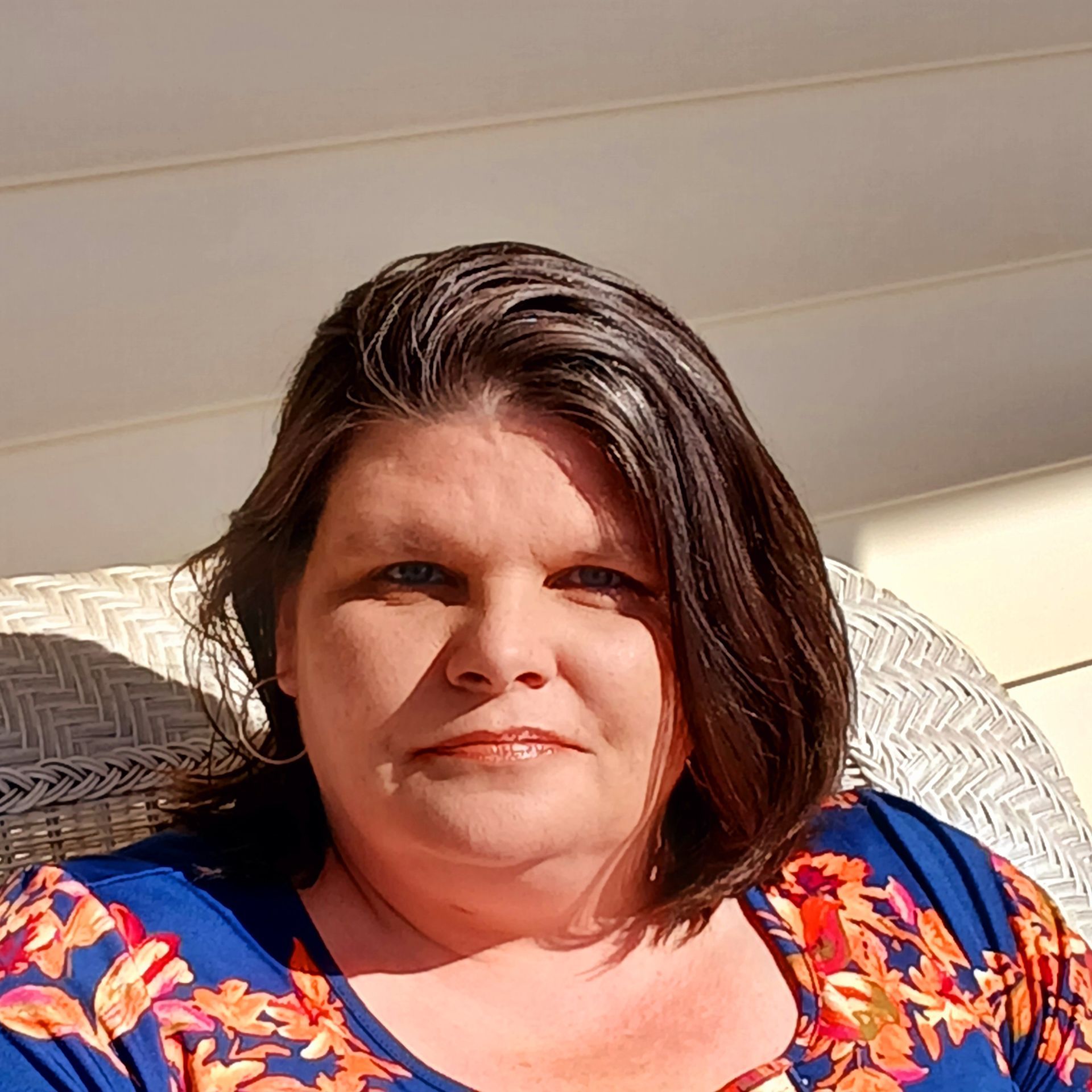 A person with dark hair wearing a blue and orange floral top sits in a wicker chair outdoors in the sunlight.