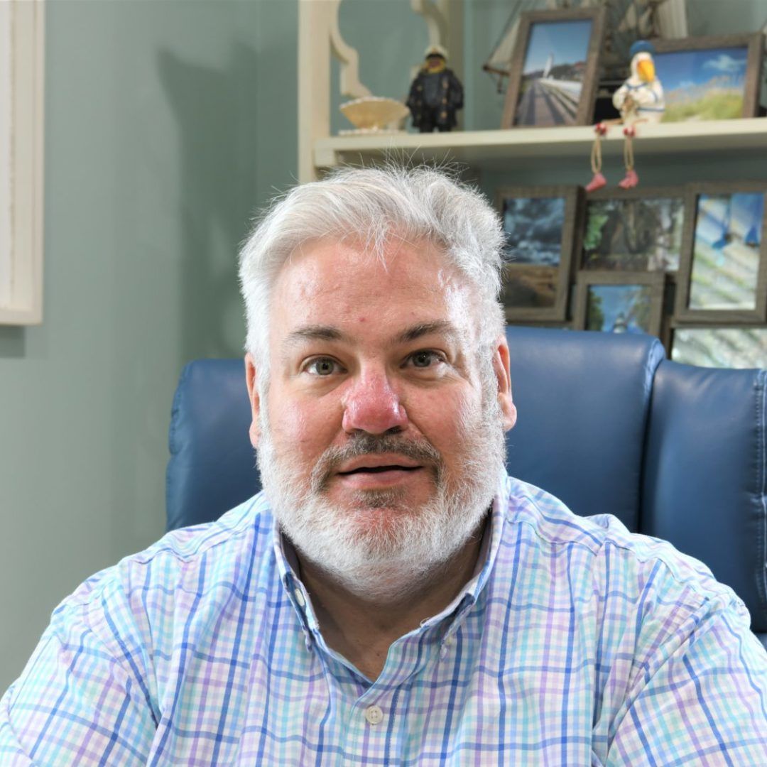 A person with white hair and a beard wearing a light blue and white checkered shirt, seated in a blue chair.