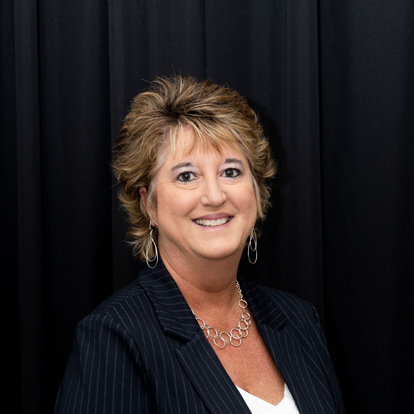 A smiling person wearing a dark pinstriped blazer, white top, and silver necklace against a black curtain background.