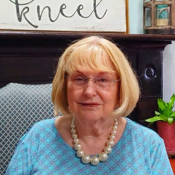 A woman with shoulder-length blonde hair and glasses wears a pearl necklace and a blue shirt, seated indoors.