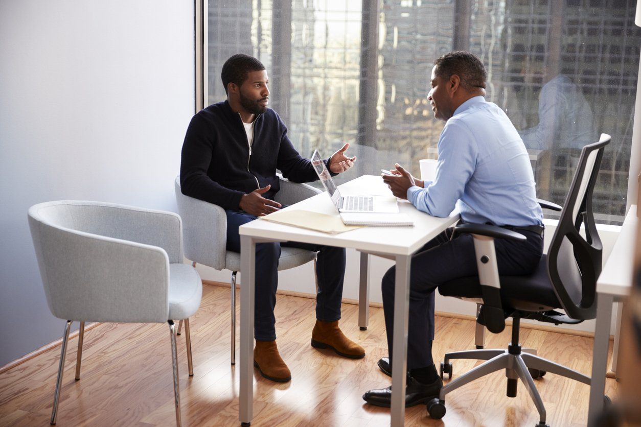 Two Black men in an office setting; one gesturing while speaking, the other seated at a desk, listening.