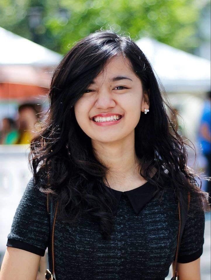 Smiling young woman with dark hair and black collared shirt outdoors.