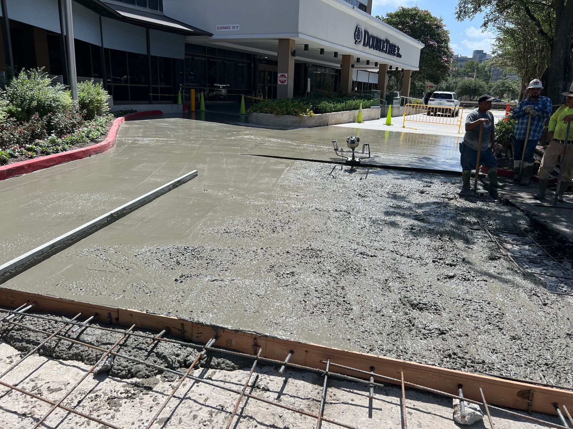 Workers pouring wet concrete sidewalk in front of a building with people observing.