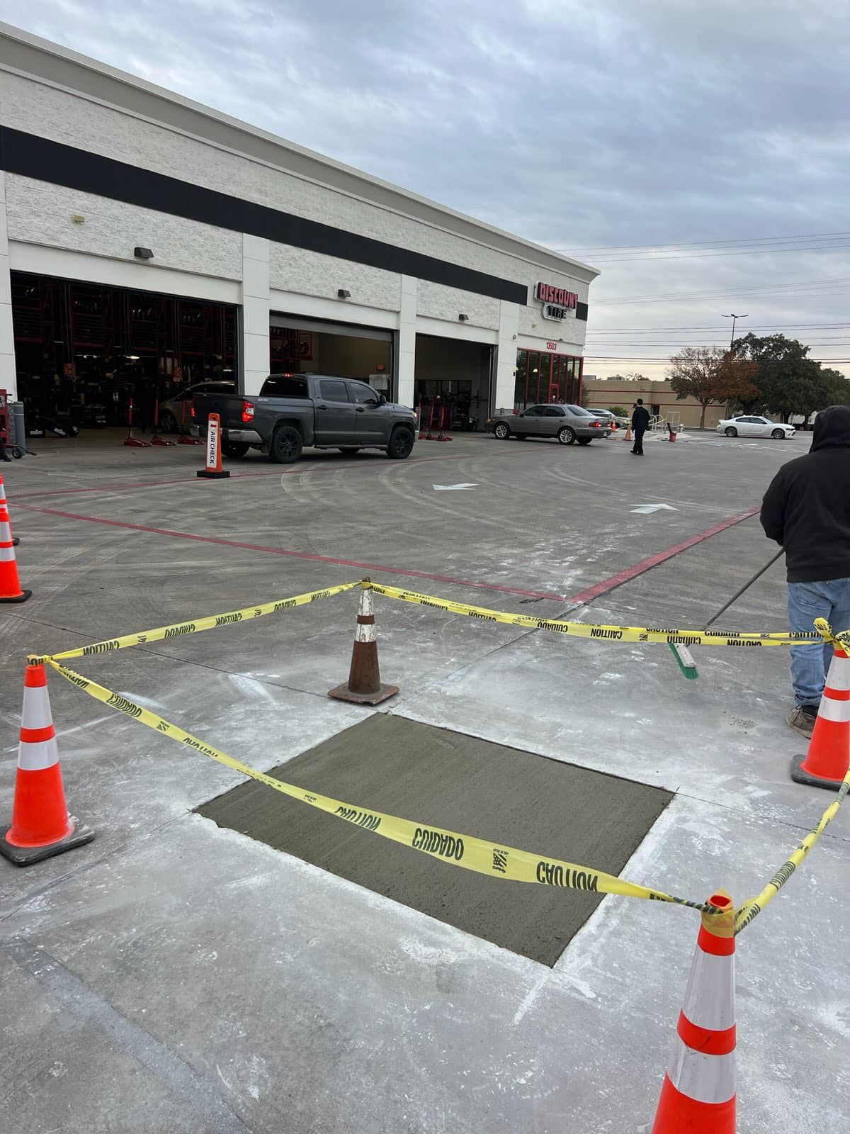 Exterior of a tire shop. Freshly poured concrete patch enclosed by caution tape, cones, and a person sweeping.