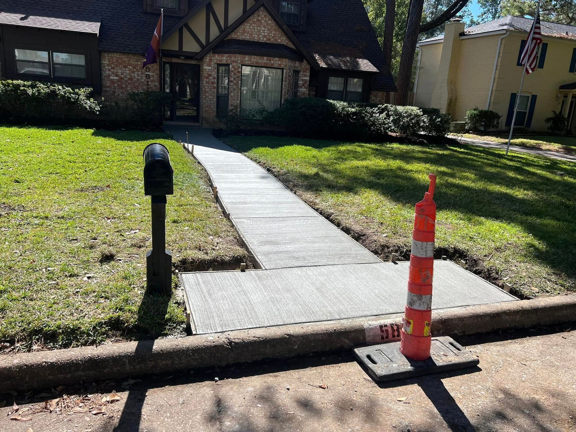A newly constructed concrete sidewalk leading to a Tudor-style home, orange cone in foreground.