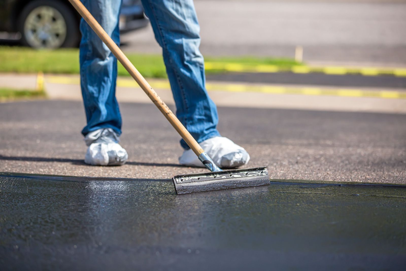 Person in jeans, shoe covers using a squeegee to apply black asphalt sealant to a driveway.
