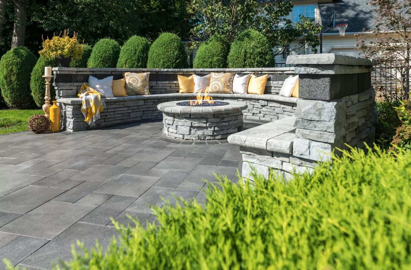 Stone patio with built-in curved bench, fire pit table, and yellow cushions in a landscaped garden