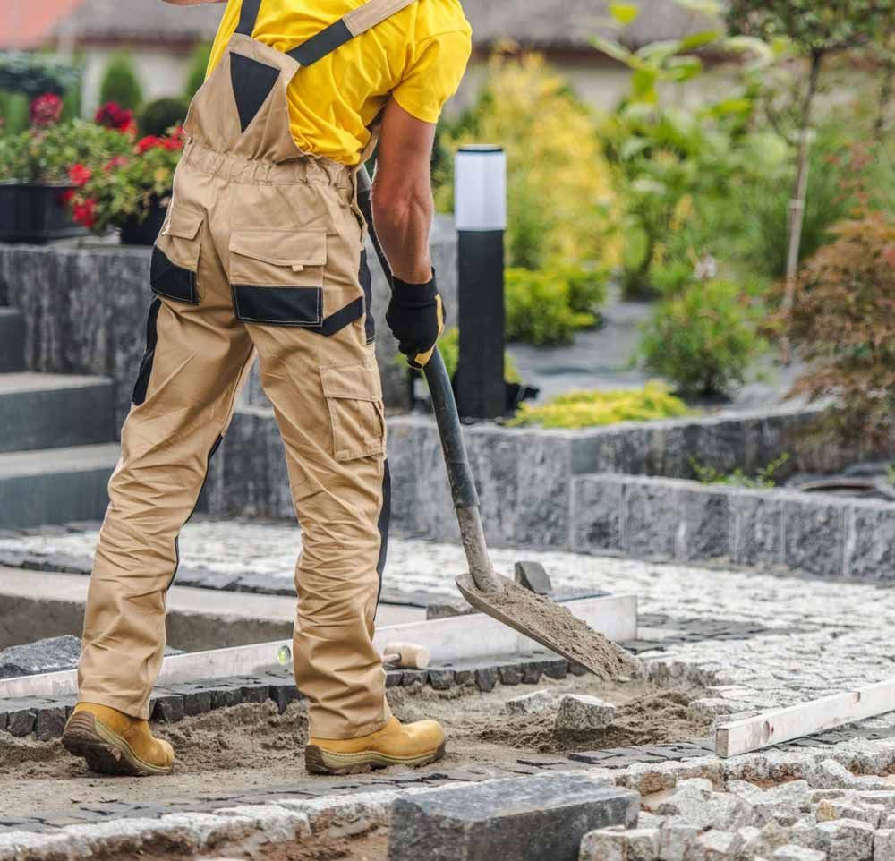 Worker in yellow shirt and tan overalls shoveling dirt on a stone walkway outdoors