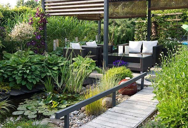 Garden patio with black seating, wooden deck, and lush greenery beside a pond