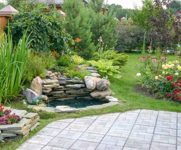 Lush backyard garden with a small rock waterfall pond, green shrubs, and a stone patio in the foreground