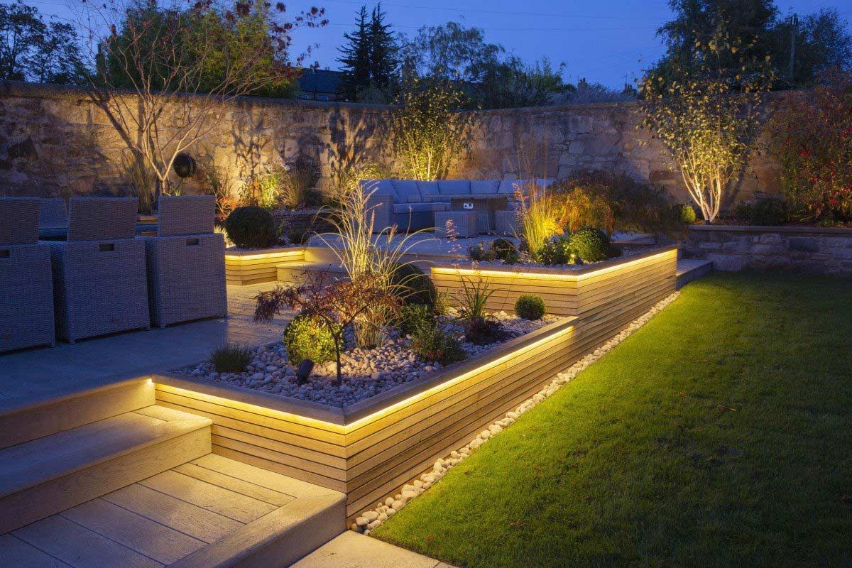 Illuminated modern backyard patio with stone seating, planters, and a green lawn at dusk