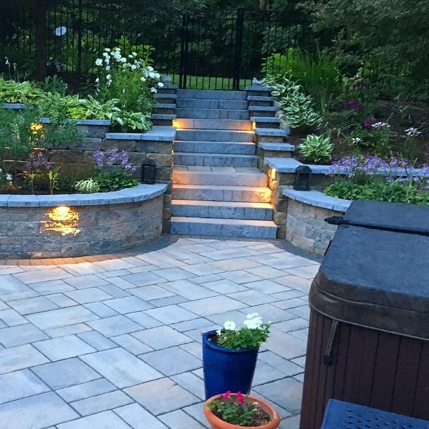 Backyard stone patio with lit steps, curved retaining walls, and potted flowers at dusk