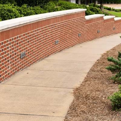 Curved concrete walkway beside a brick retaining wall with greenery above and mulch along the edge