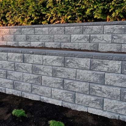 Stone retaining wall with gray block tiers and dark mulch at the base, bordered by greenery above.