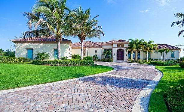 Luxury single-story house with palm trees, manicured lawn, and a curved brick driveway under a blue sky