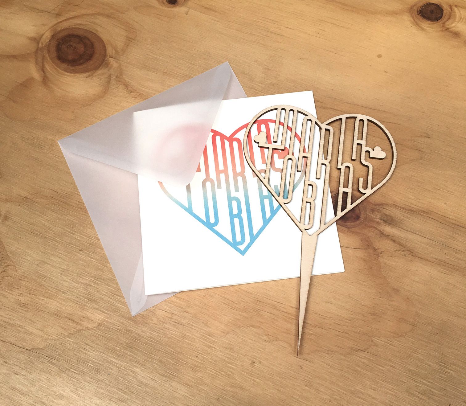 Cake-Topper aus feinem Cut-Holz mit einer Karte mit dem selben Motiv in rotblauem Verlauf.