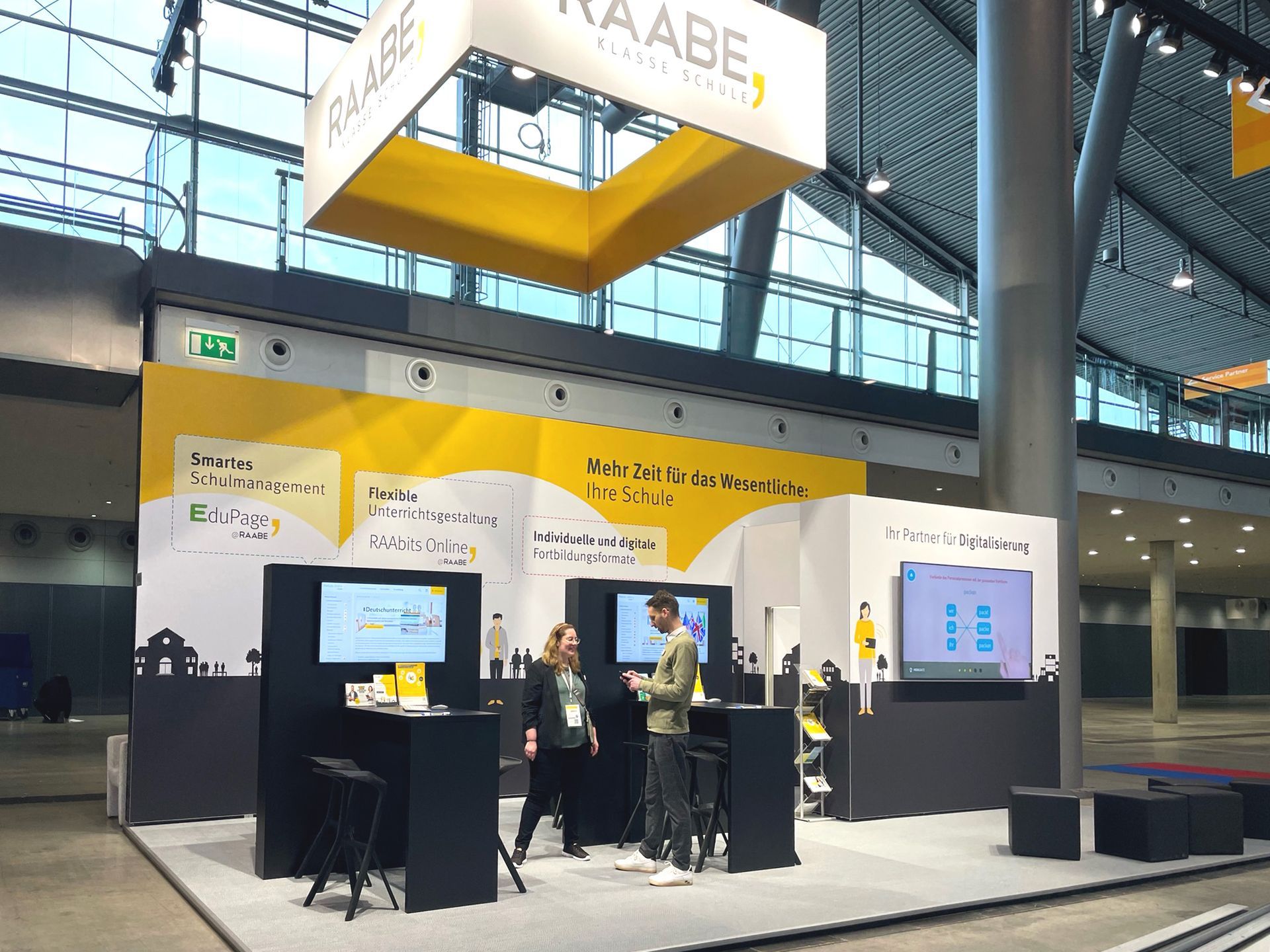 RAABE-Messestand auf der Didacta 2023 in Stuttgart Großer Messestand in RAABE-Corporate-Design, etwa 3 Meter hoch mit einer großflächigen Wand mit Illustrationen und Info-Blasen. Davor stehen zwei Tische mit Monitoren und ein Quadrat mit einem größeren Monitor. Oben drüber hängt ein großer Würfel mit dem RAABE-Logo.