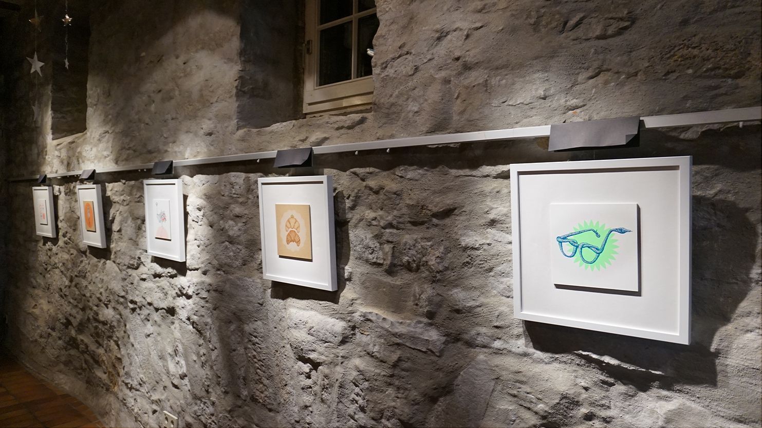 5 Illustrationen (Wortbilder) hängen in quadratischen weißen Bilderrahmen an einer rustikalen Steinwand und sind schön beleuchtet.