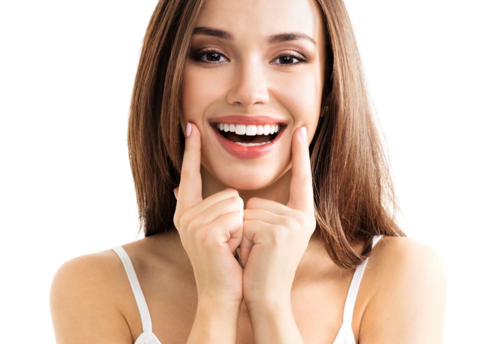 Donna sorridente, dita che indicano le guance, canotta bianca, capelli castani, sfondo bianco.