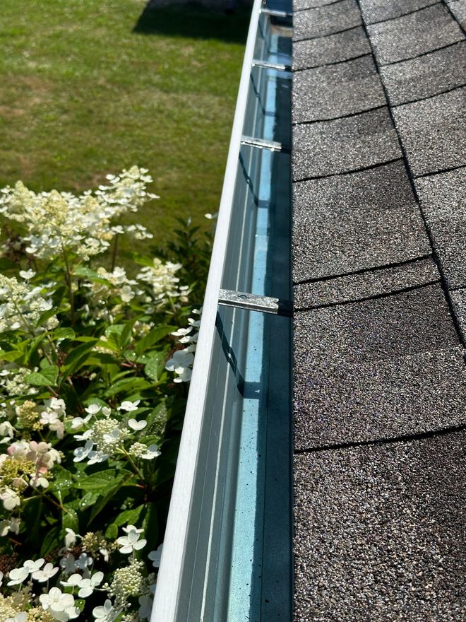 Roof gutter beside shingled roof with white flowering shrubs below and lawn in the background